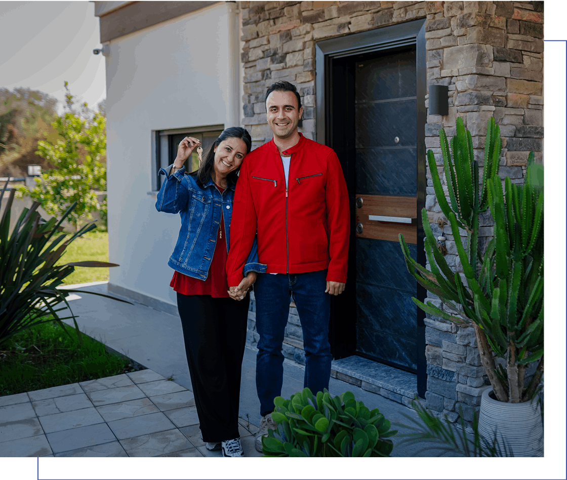 A smiling couple stands in front of a modern house, holding keys and each other's hand. They are surrounded by greenery and a stone facade.