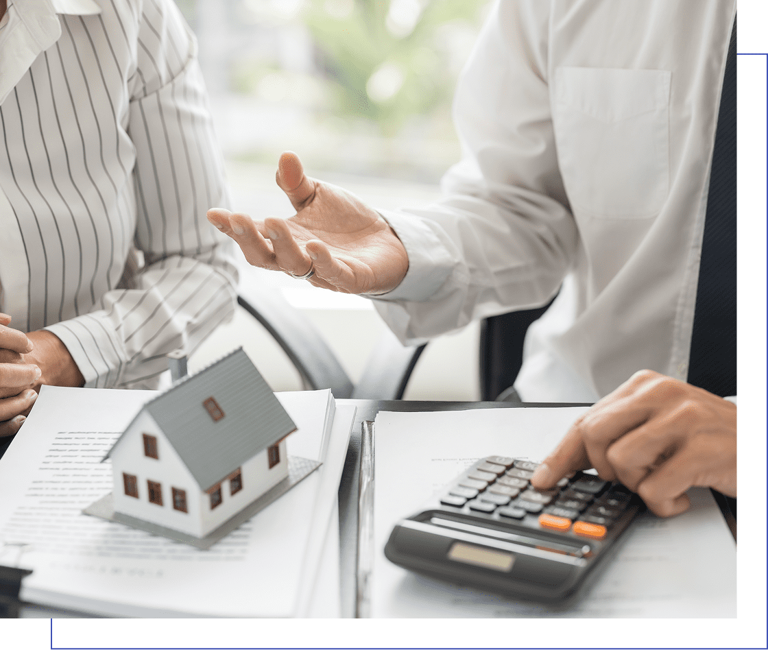 Two people in business attire discuss over paperwork with a small house model and a calculator on a desk.