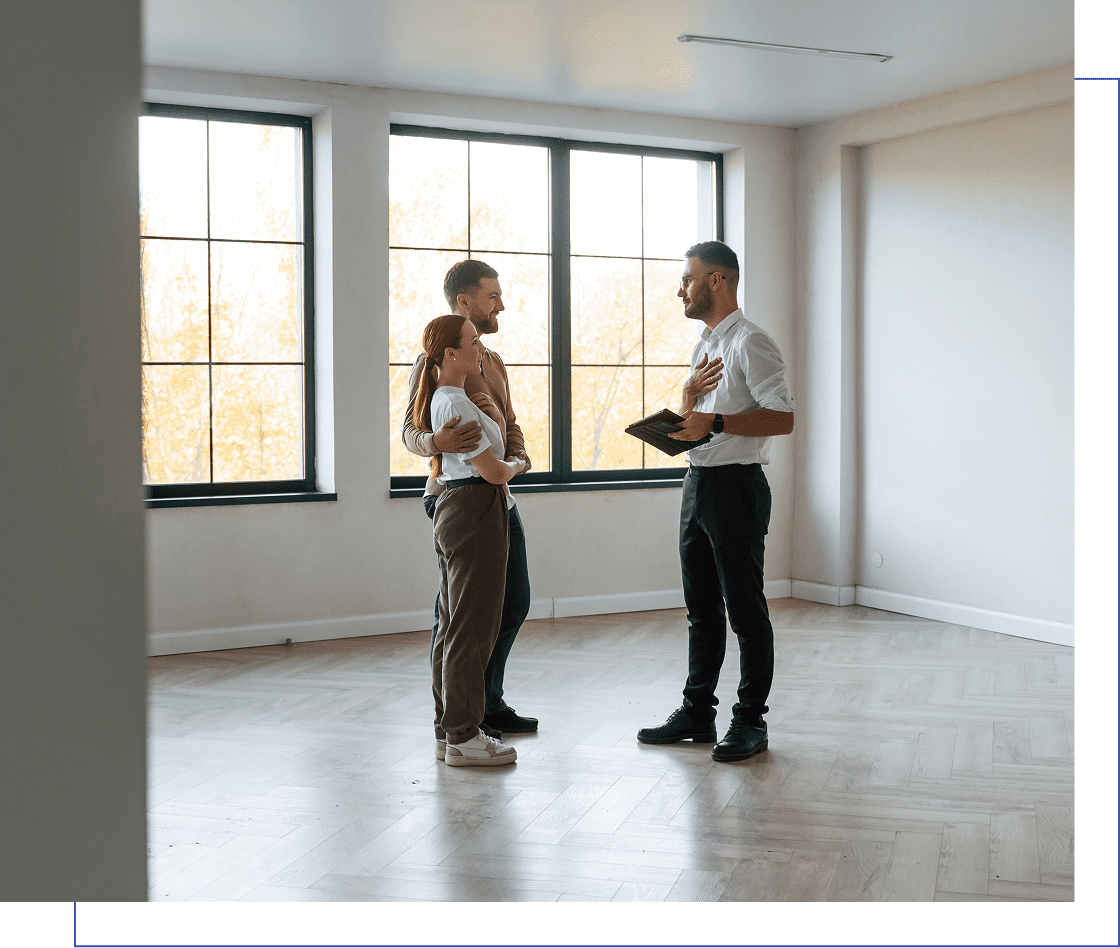 A couple holding a baby talks to a man with a clipboard in a bright, empty room with large windows.