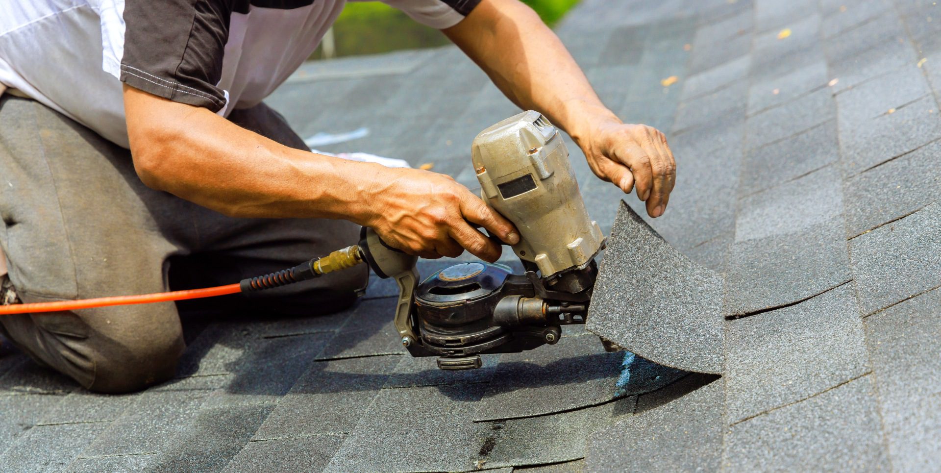 A person uses a pneumatic nail gun to install asphalt shingles on a roof.