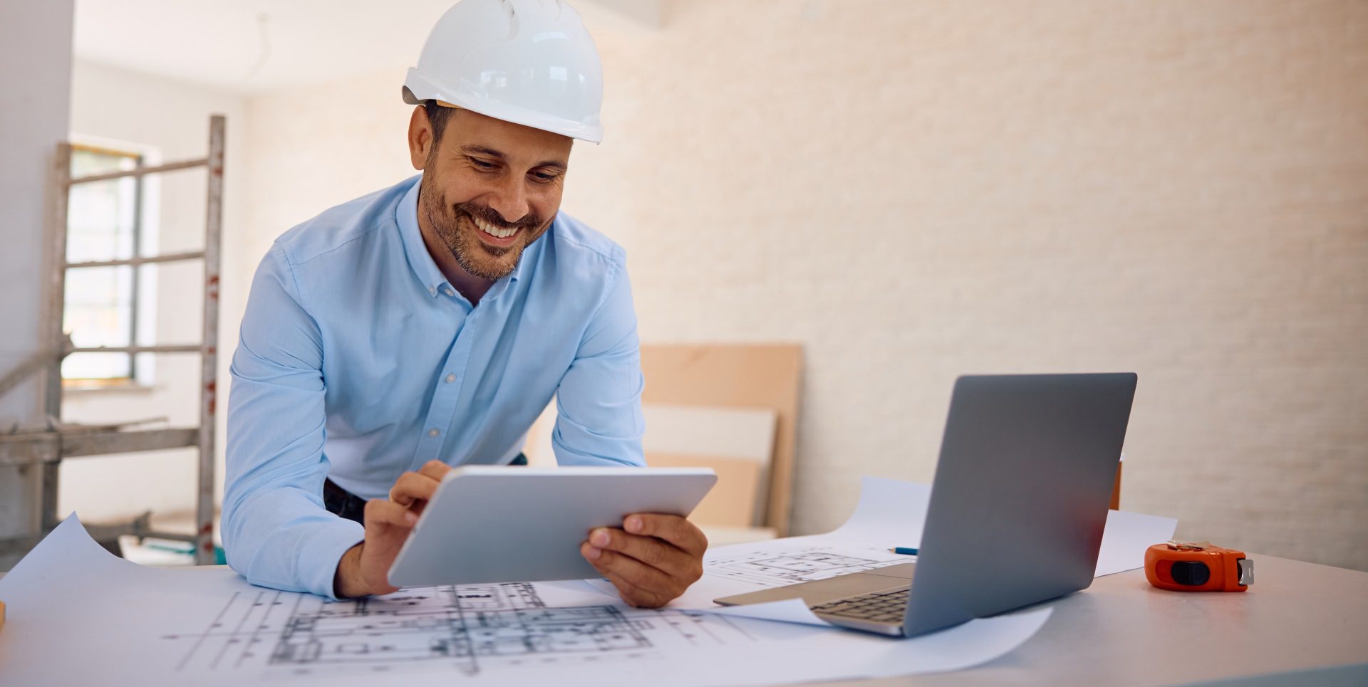 A man in a hard hat smiles while looking at a tablet, standing over architectural plans on a desk with a laptop nearby.