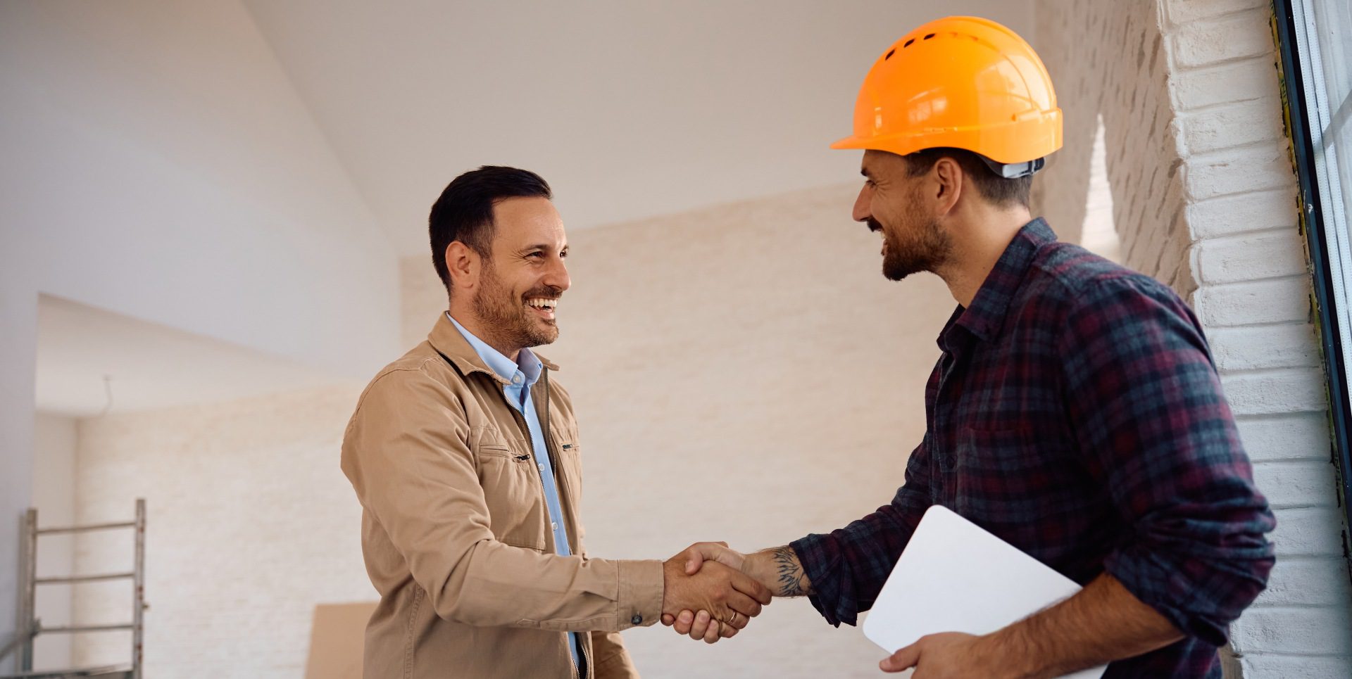 Two men smiling and shaking hands indoors. One wears a construction helmet and holds a tablet.