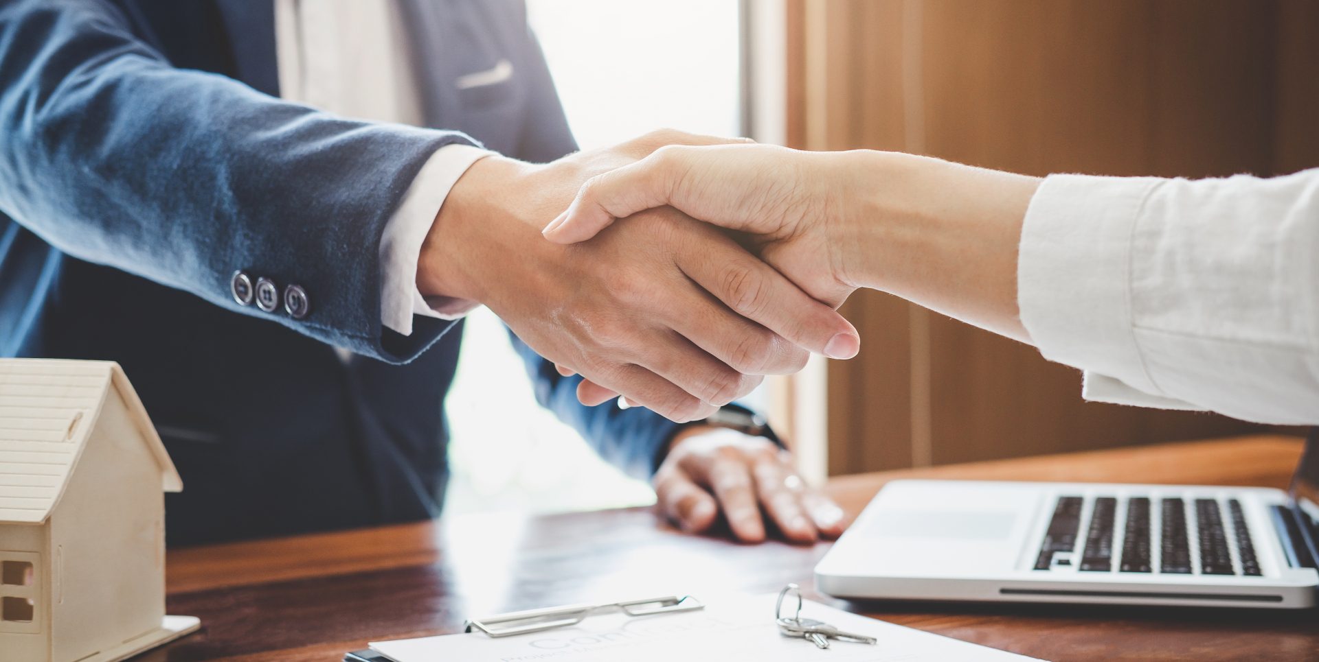 Two people shaking hands over a desk with a laptop, small house model, and documents.